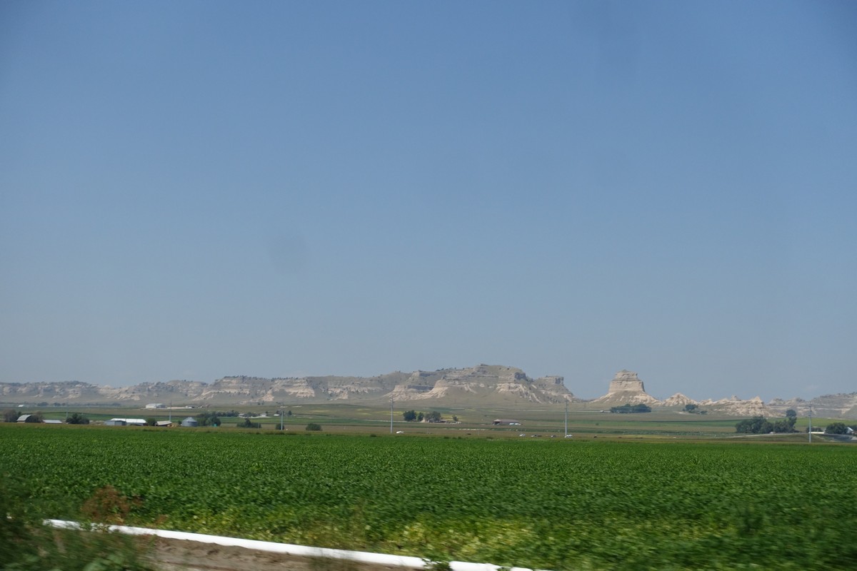 Nebraska landscape with green fields, distant buttes, and scattered farm buildings under a clear sky.