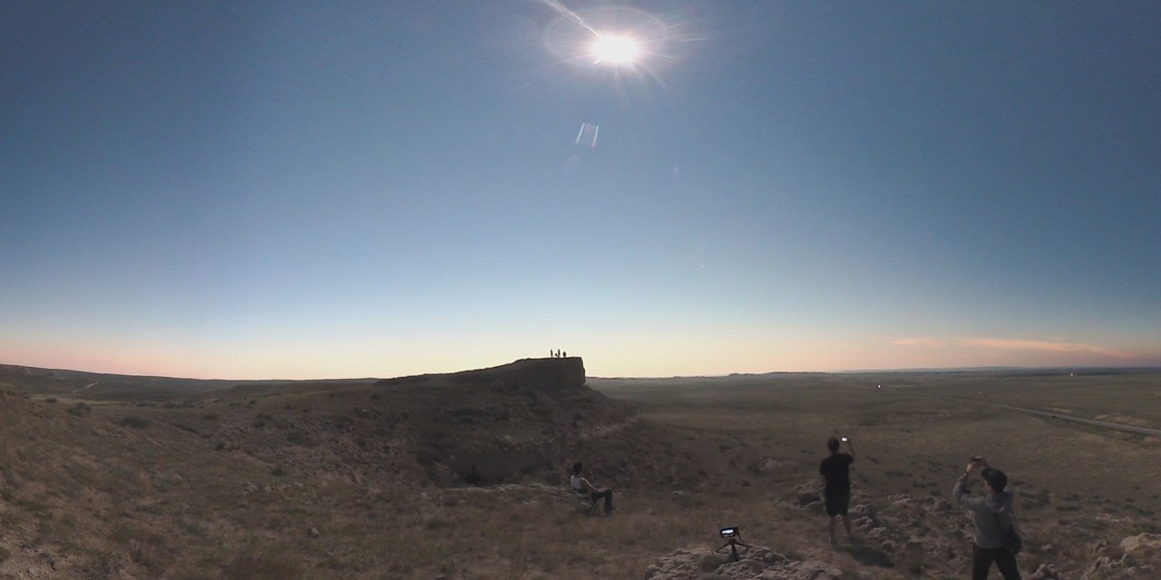 A bright sun shines over a vast Nebraska landscape where people are gathered to watch an eclipse.