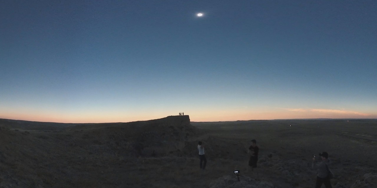 A group of people watch a total solar eclipse from a dark, rugged landscape in Nebraska.