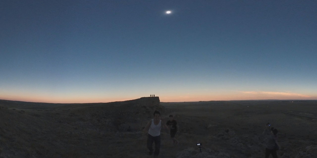 A brilliant total solar eclipse is visible in the darkening sky above a vast, open landscape with people observing.