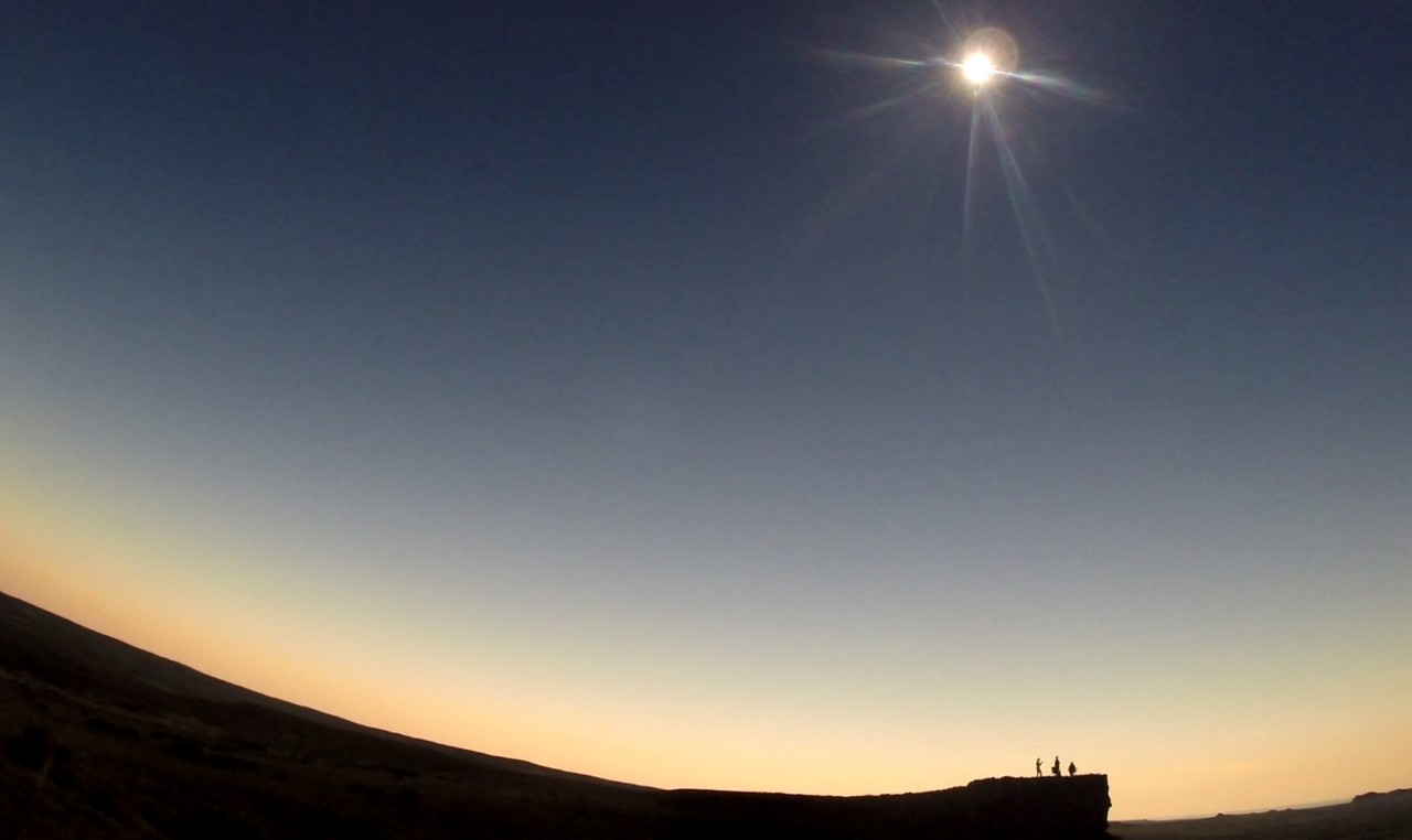 A group of people are silhouetted on a bluff against a gradient sky, watching a solar eclipse with prominent sun rays.