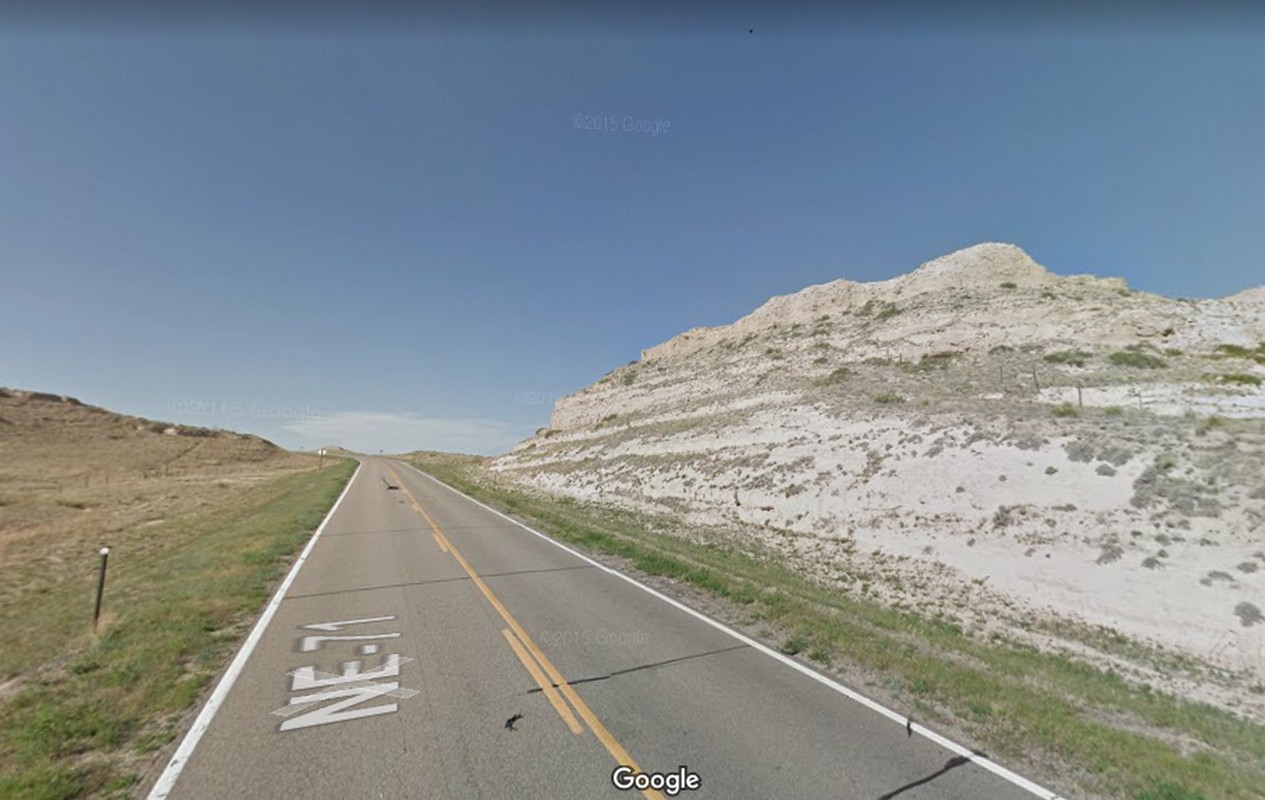 A paved road with "NE-71" painted on it passes dry, grassy hills and a large rocky bluff under a clear blue sky.