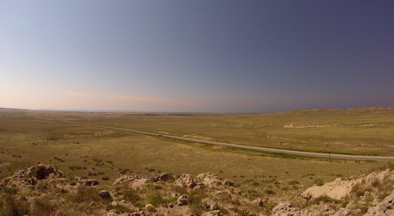 A long road extends through vast grassy plains with a rocky foreground under a clear sky in Nebraska.