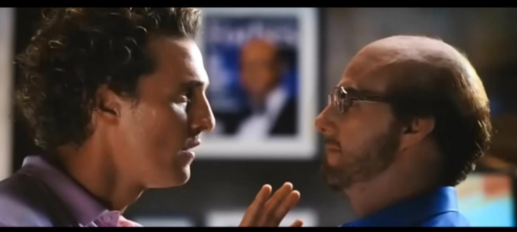 Two men, one with curly hair and a purple shirt, the other balding with glasses and a beard, face each other closely.