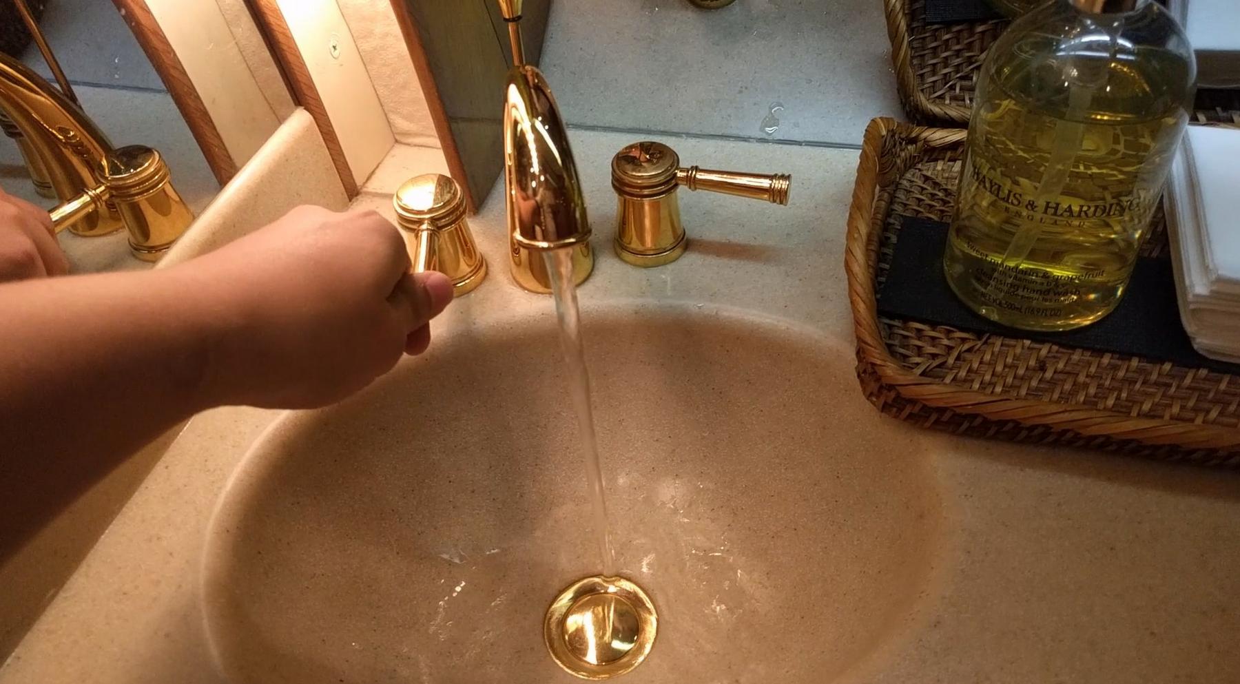 A hand turns on a golden faucet with water flowing into a sink in a luxurious private jet lavatory.