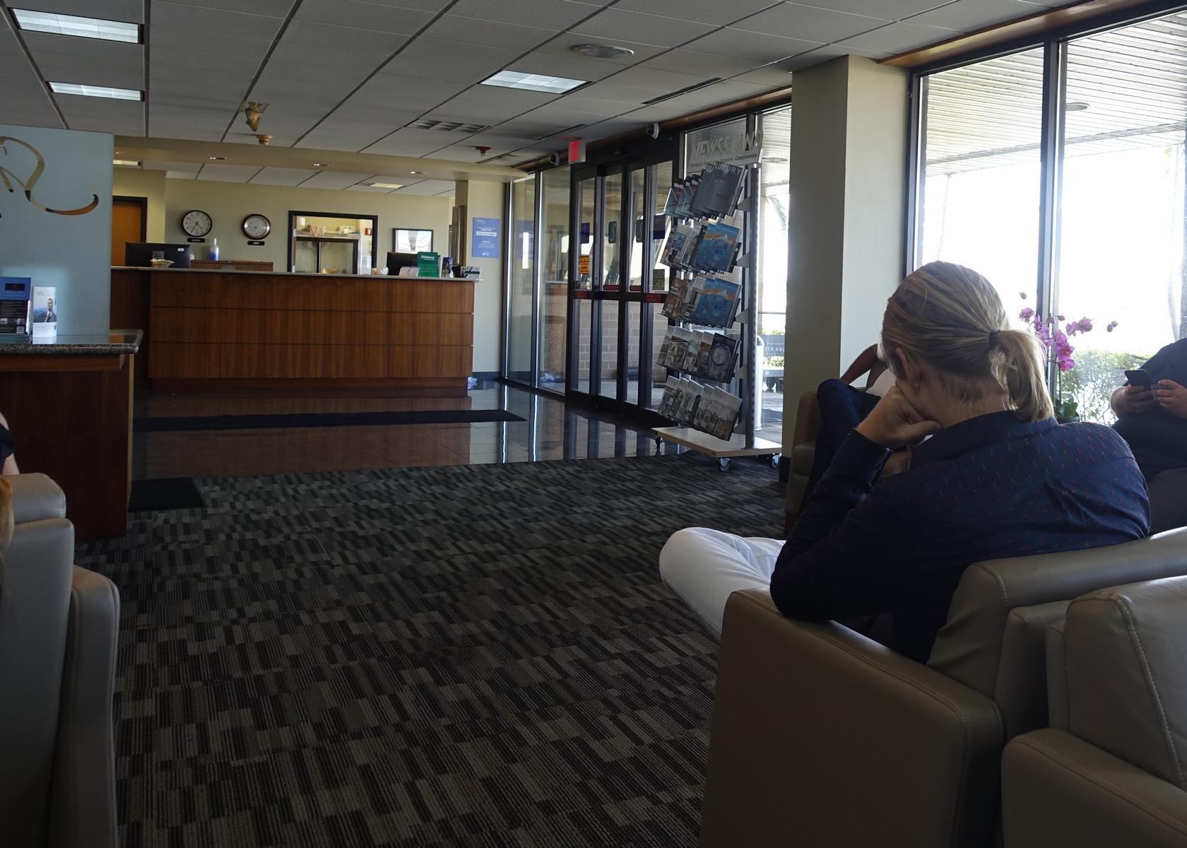 A person sits in a private jet terminal lounge with a reception desk, patterned carpet, and large windows looking outside.