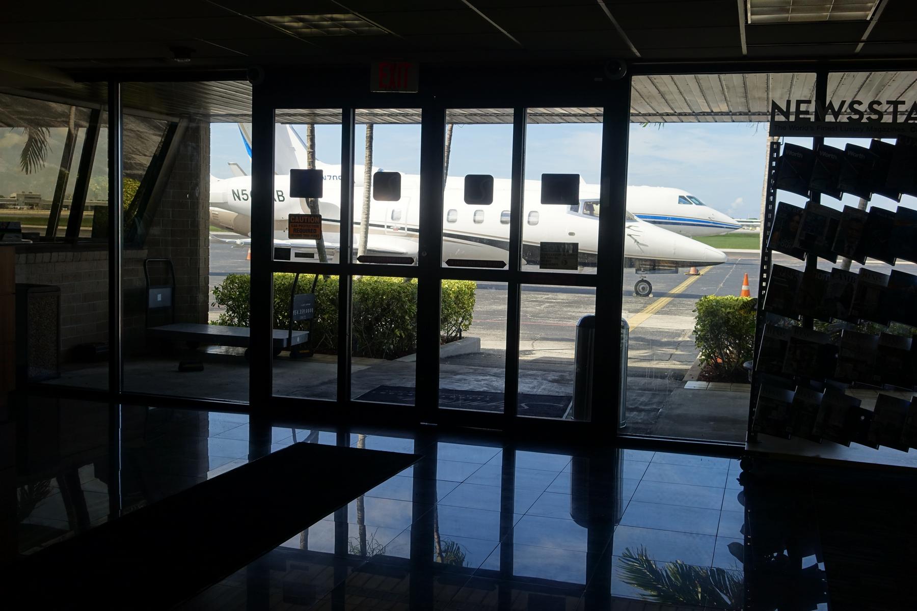 From a dark private jet terminal, a view through glass doors shows two private jets on a sunny tarmac.