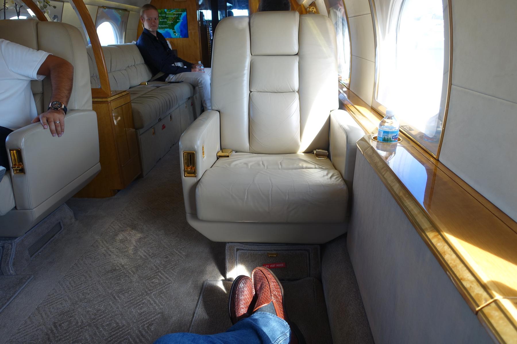 First-person view inside a luxurious private jet cabin with cream leather seats, wood accents, and a passenger in the background.