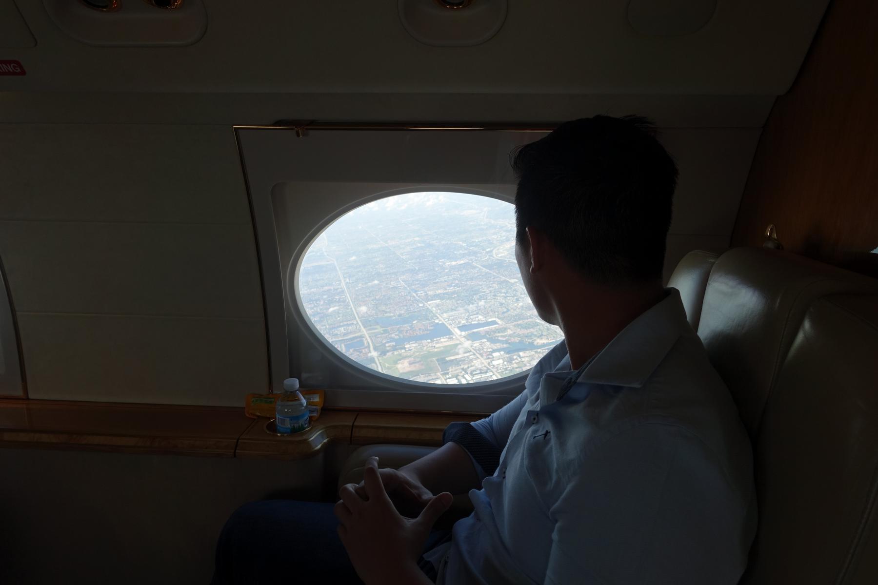 A man looks out the oval window of a private jet at the city below.