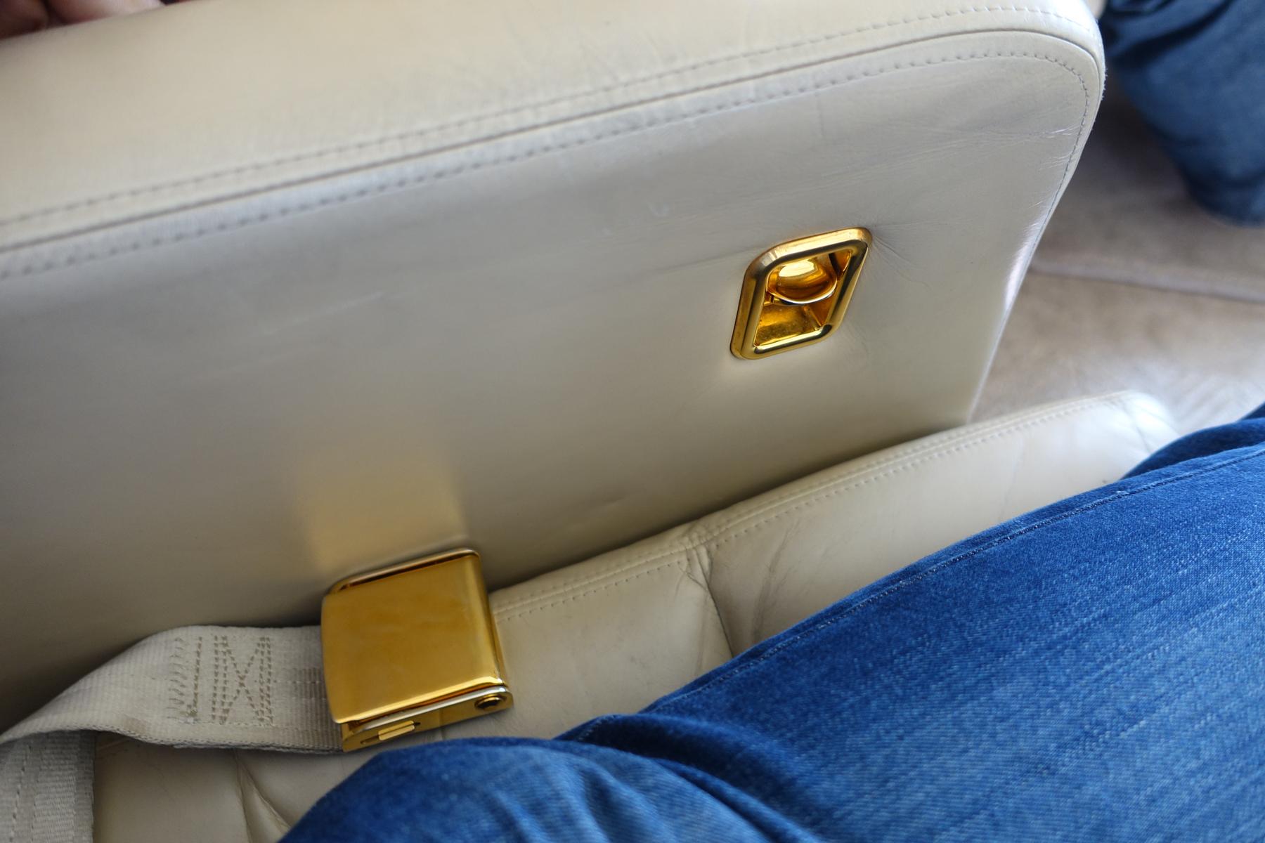 Close-up of a beige leather private jet seat with gold accents and a gold seatbelt buckle, next to a person wearing blue jeans.