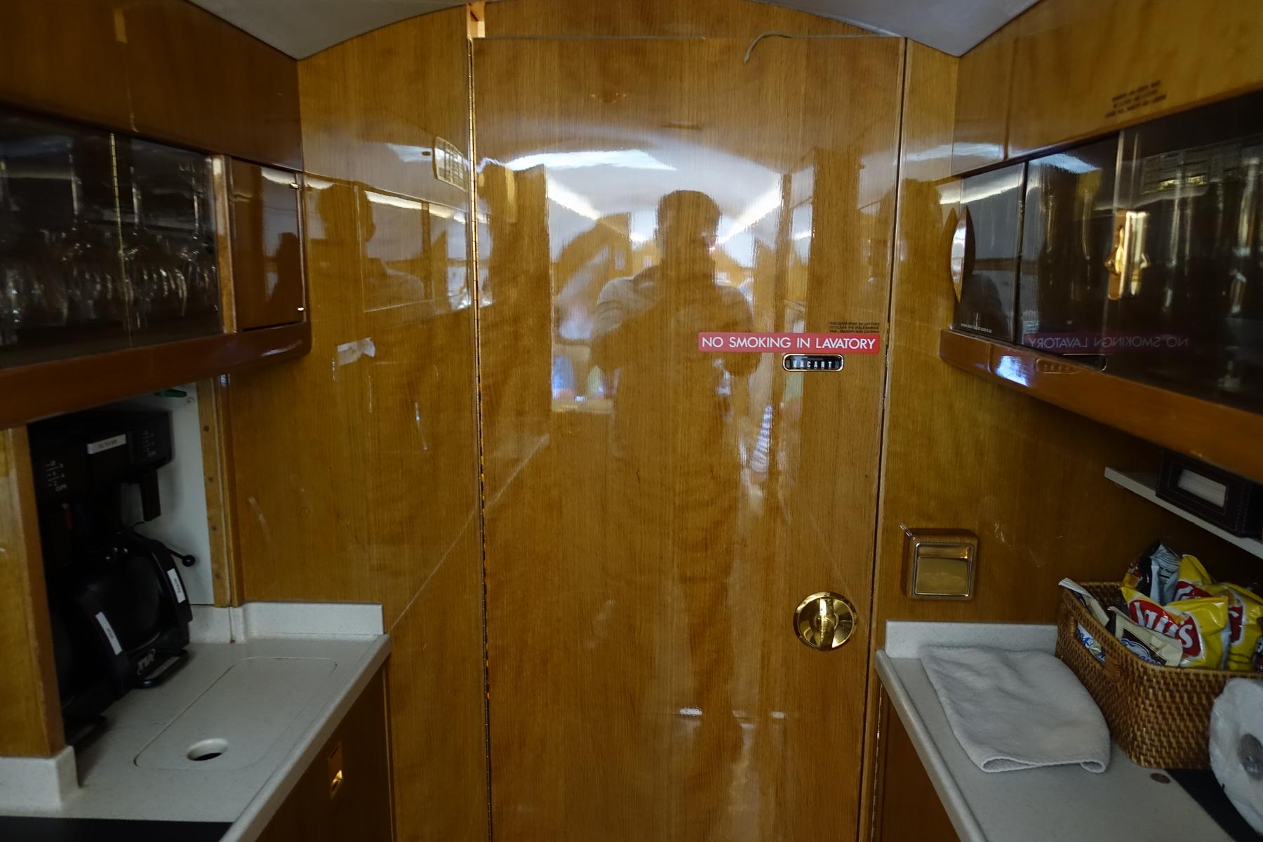 The polished wood interior of a private jet's galley area, showing a lavatory door, counters with drinks and snacks, and reflections of the cabin.