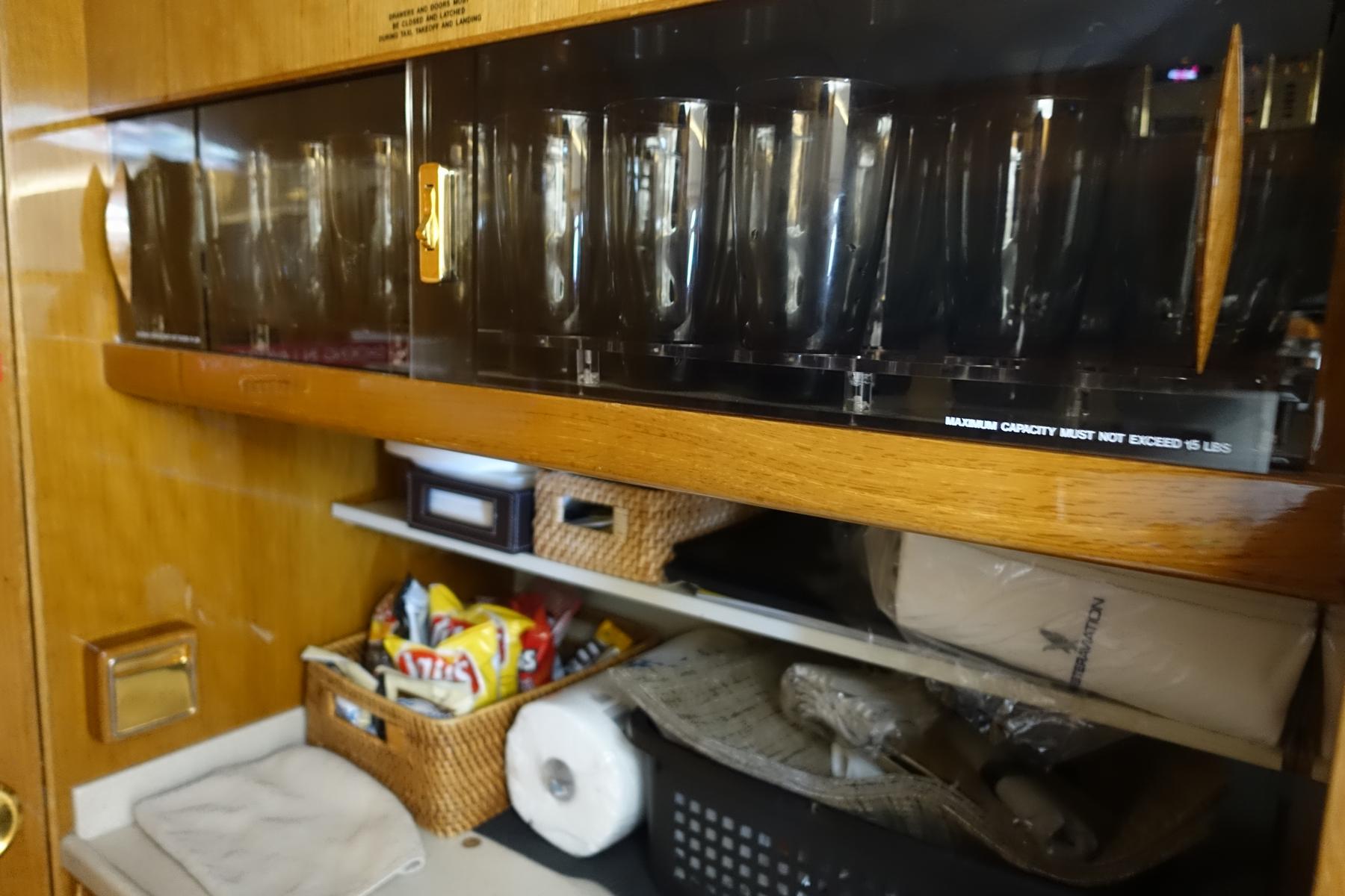 A private jet's galley area features a secured rack of clear glasses above shelves holding snacks and supplies.