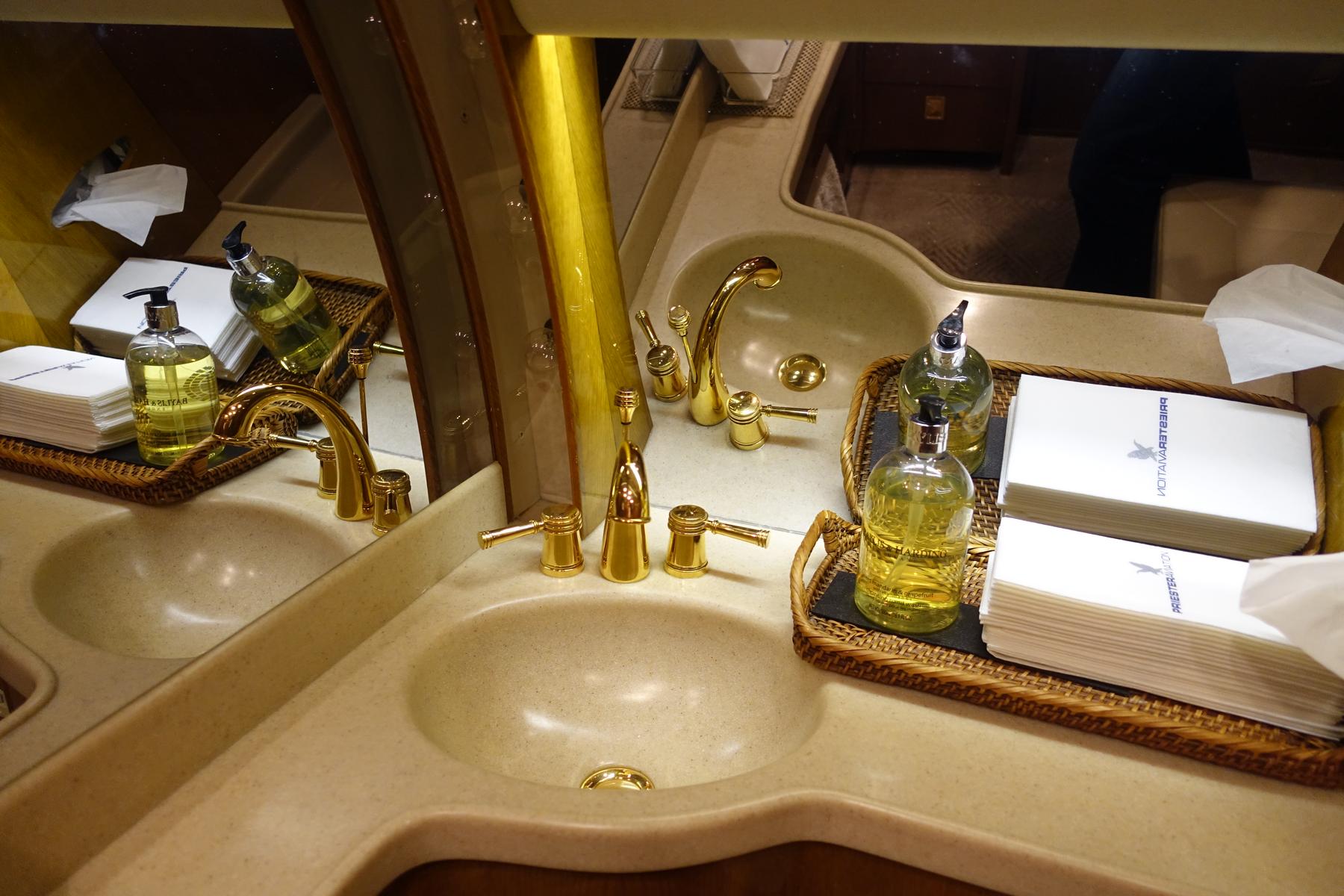A private jet lavatory featuring dual sinks with gold faucets and Molton Brown toiletries.