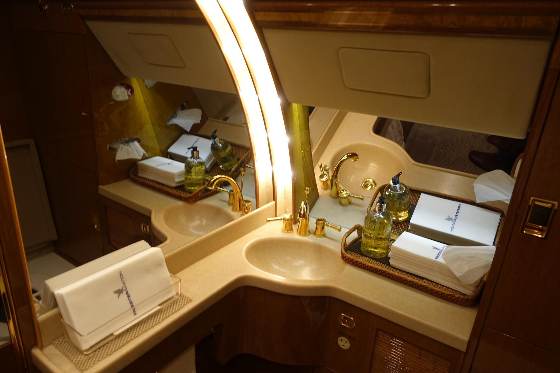 A luxurious private jet lavatory features dual sinks with golden faucets, wood paneling, and neatly arranged bottled soap and folded towels.