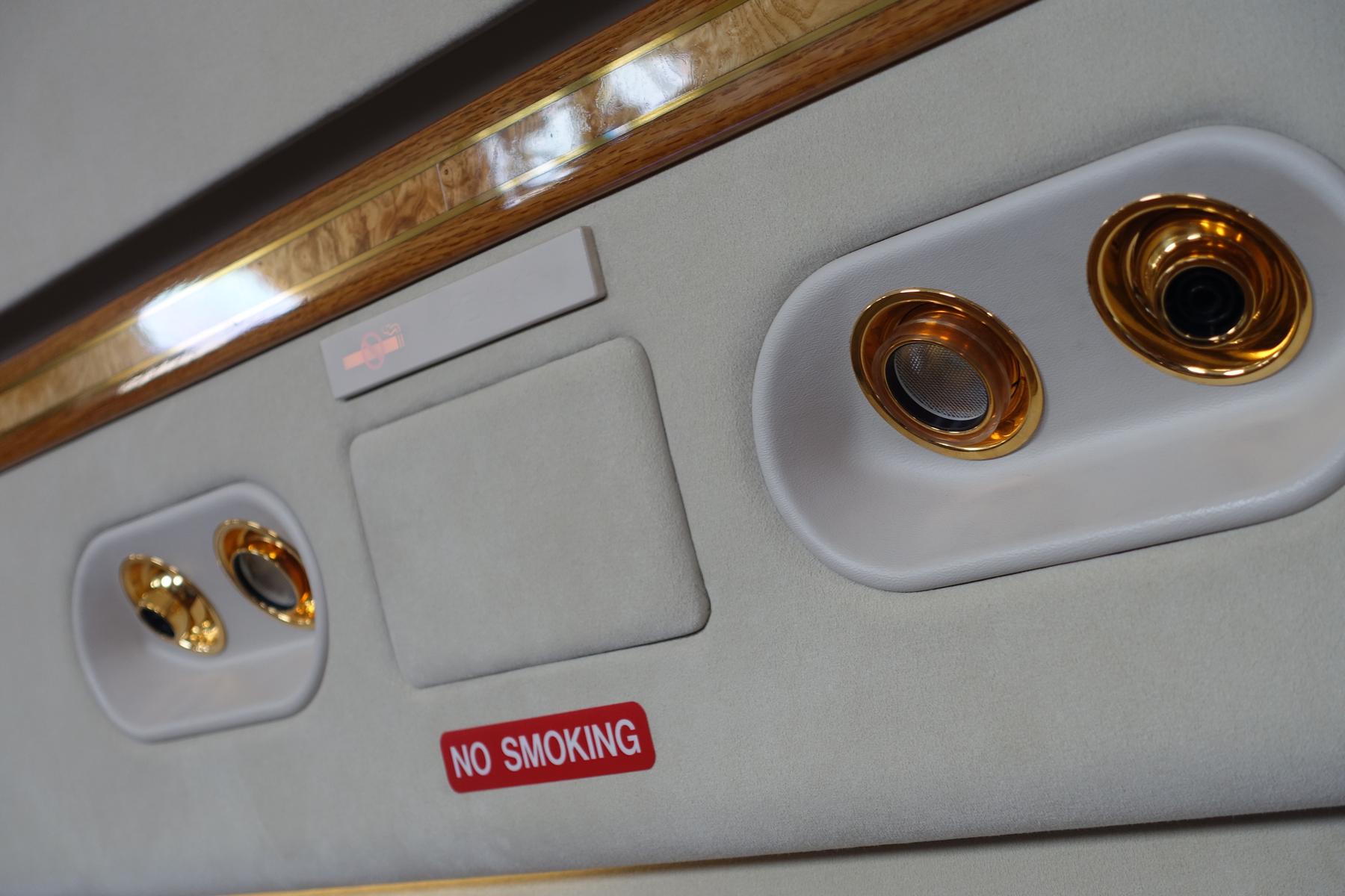 Interior view of a luxurious private jet's overhead panel with wood trim, gold accents, and a "NO SMOKING" sign.