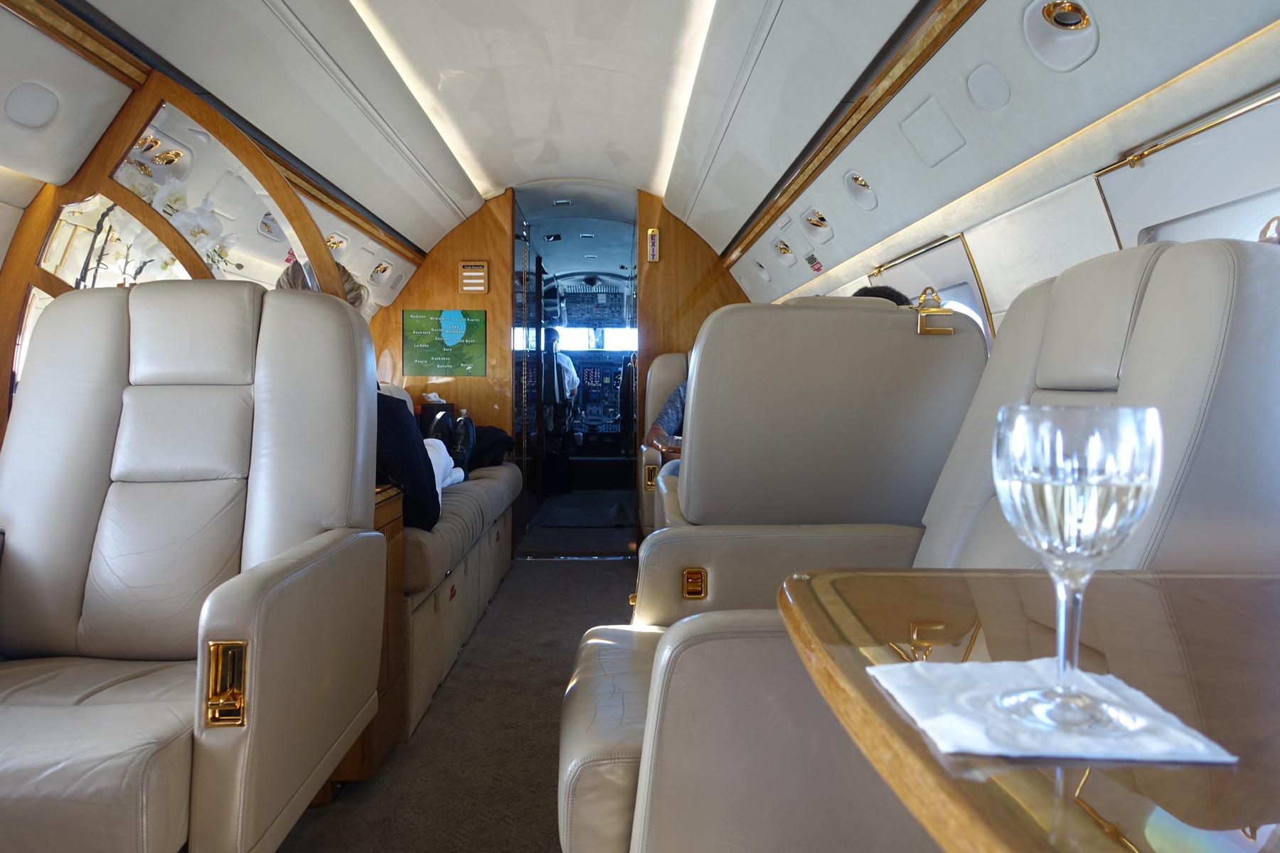 The luxurious interior of a Gulfstream G5 private jet, featuring cream leather seats, wood trim, a glass of white wine, and a view into the cockpit.