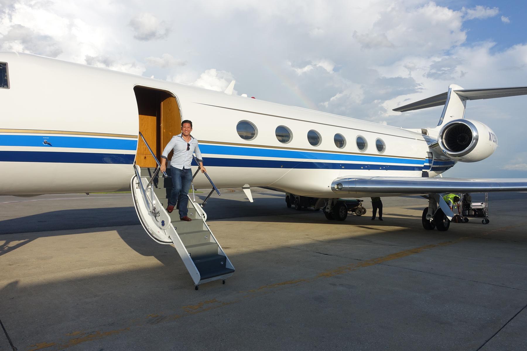 A smiling man descends the airstairs from a white Gulfstream G5 private jet, with a faint rainbow visible in the sky.