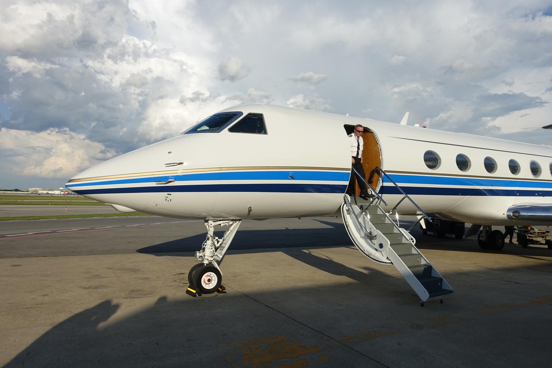 A pilot stands in the open doorway of a white Gulfstream G500 private jet with blue and gold stripes on the tarmac.