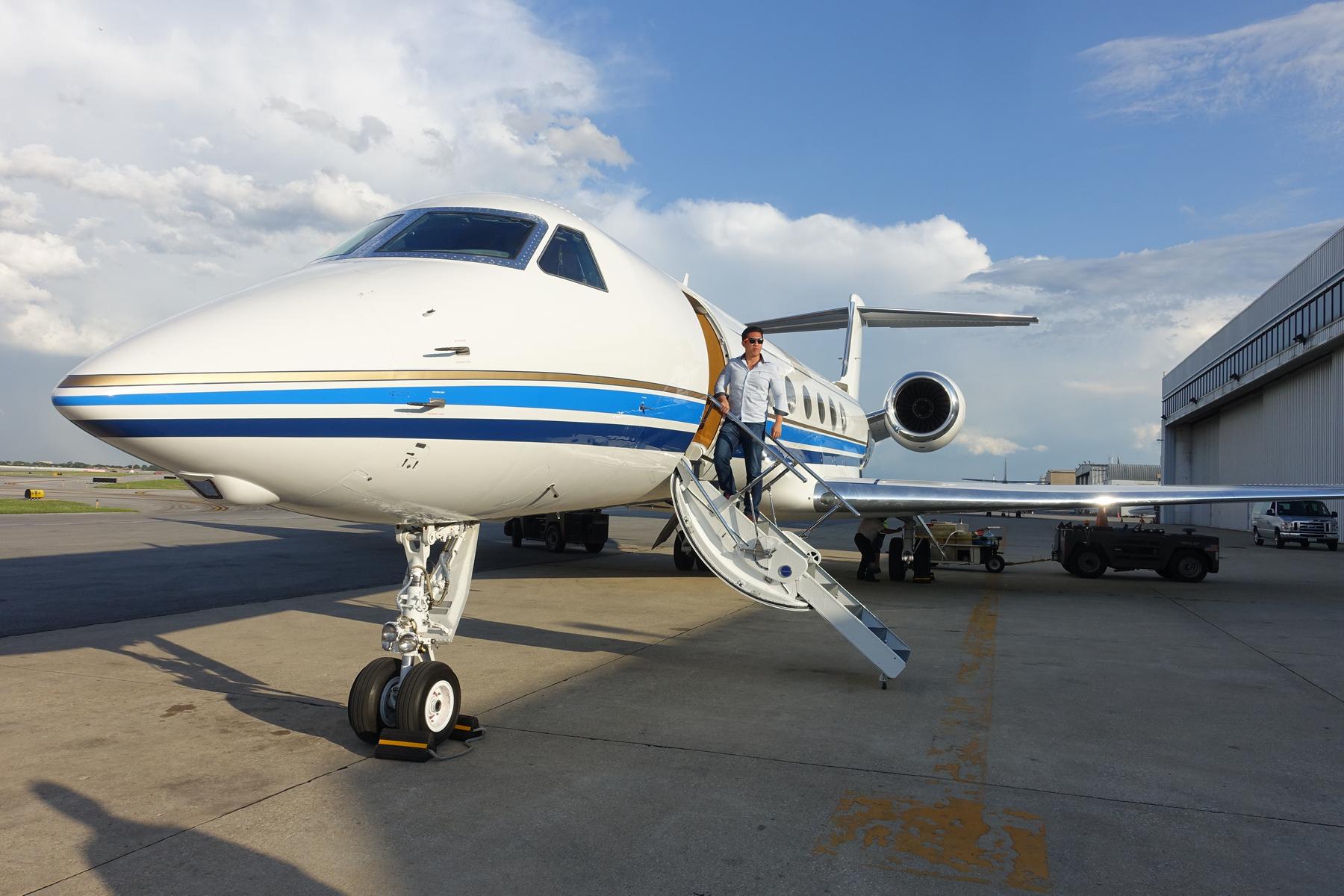A man descends the stairs of a white Gulfstream 500 private jet with blue and gold stripes on an airport tarmac.