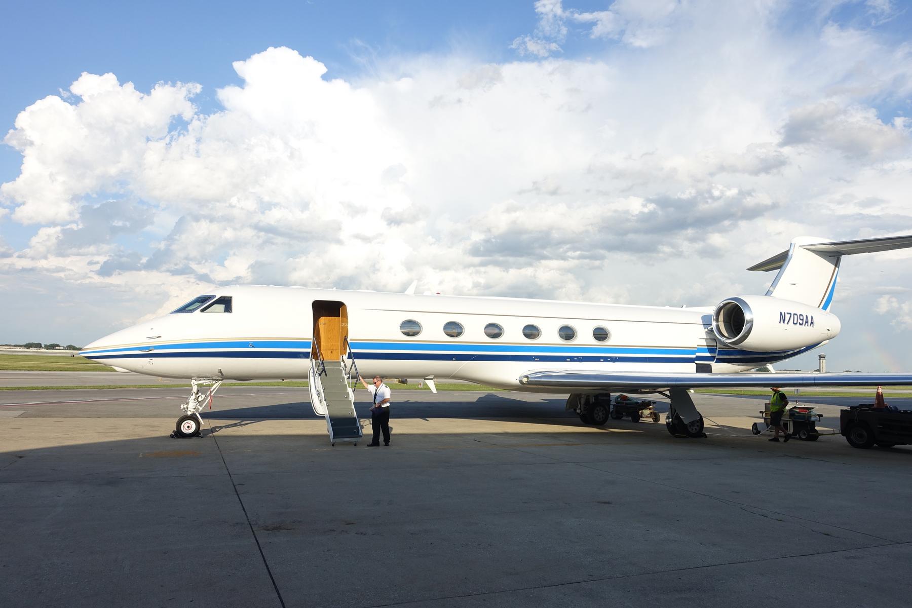 A white and blue Gulfstream 500 private jet with open stairs on the tarmac, with a pilot standing by.