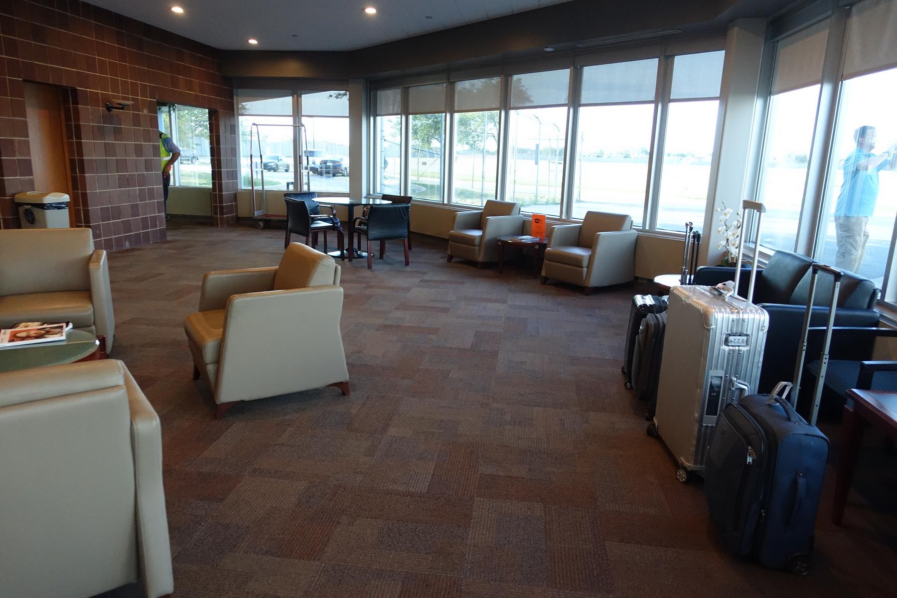 A lounge area in a private jet terminal with armchairs, tables, luggage, and large windows.