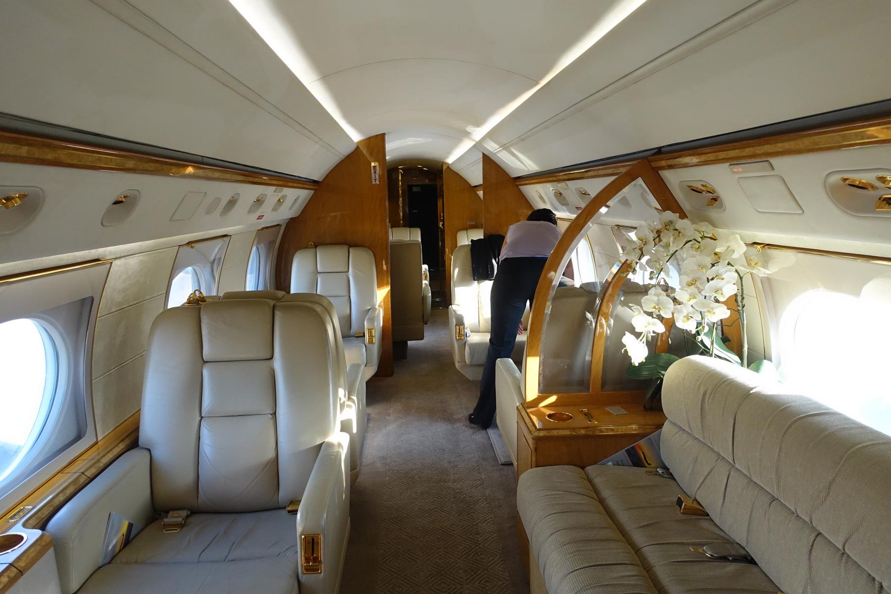 A person stands in the luxurious cabin of a private jet, featuring beige leather seats, wood accents, and white orchids.