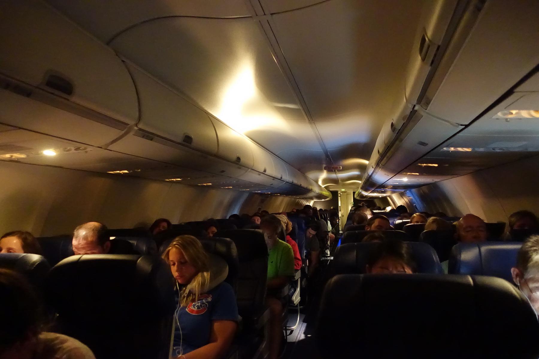 A dimly lit, crowded commercial airplane cabin with rows of seated passengers.