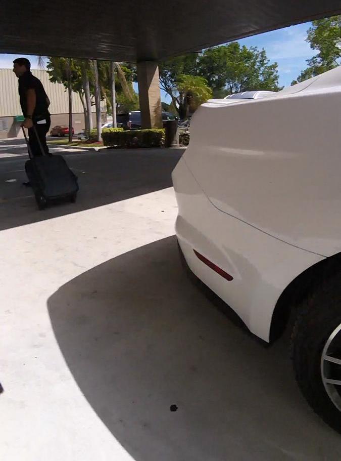 A man pulls a suitcase past a white car under a covered walkway.