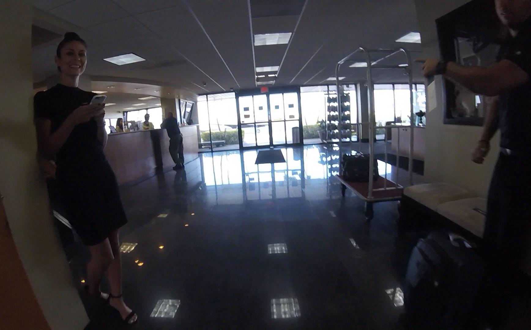 A smiling woman holding a phone stands in a bright hotel lobby next to a luggage cart.