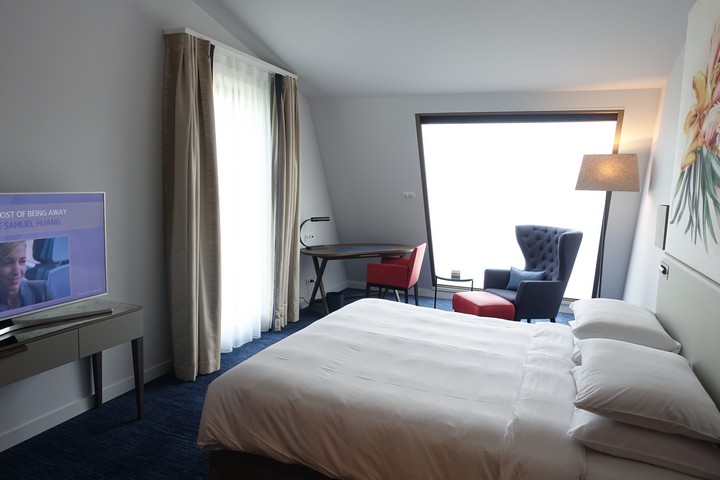 A modern hotel room with a white bed, blue carpet, TV, a desk with a red chair, and a blue armchair with a red ottoman next to a bright window.