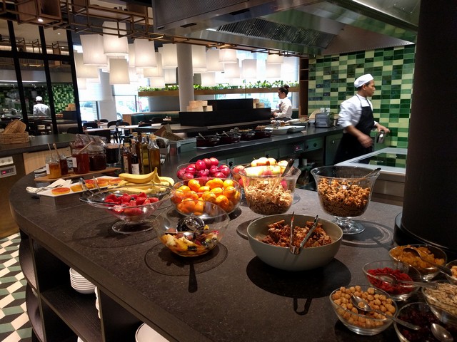 A modern hotel breakfast buffet features bowls of fresh fruit, granola, nuts, and preserves, with kitchen staff visible in the background.