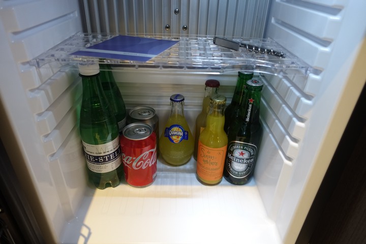 A hotel minibar is filled with assorted bottled drinks, cans, and a corkscrew on the shelf.