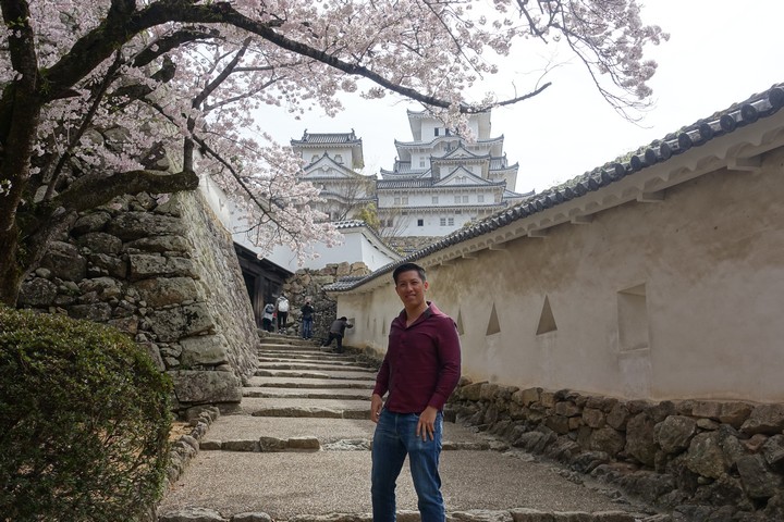 A smiling man stands on a stone path lined with cherry blossoms, with Himeji Castle towering in the background.