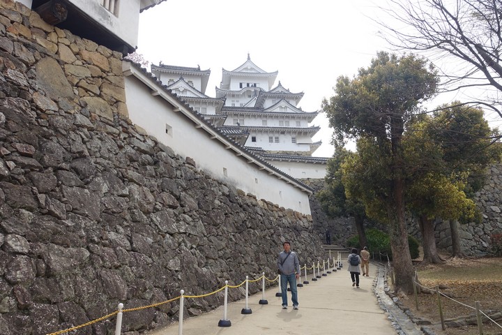Himeji Castle's white multi-tiered keep towers over extensive stone walls, with a path leading to it and visitors walking.