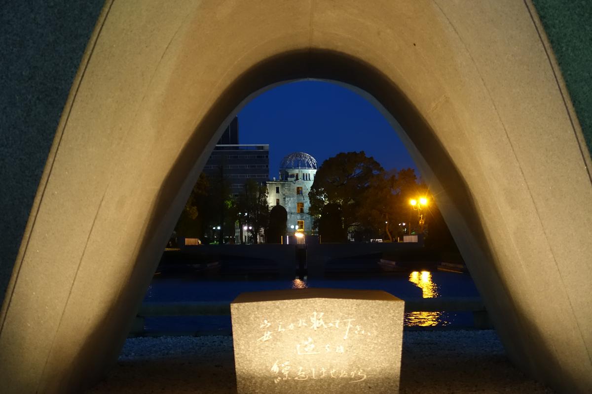 The Atomic Bomb Dome, lit at night, framed by the Memorial Cenotaph arch and a stone monument in Hiroshima Peace Park.