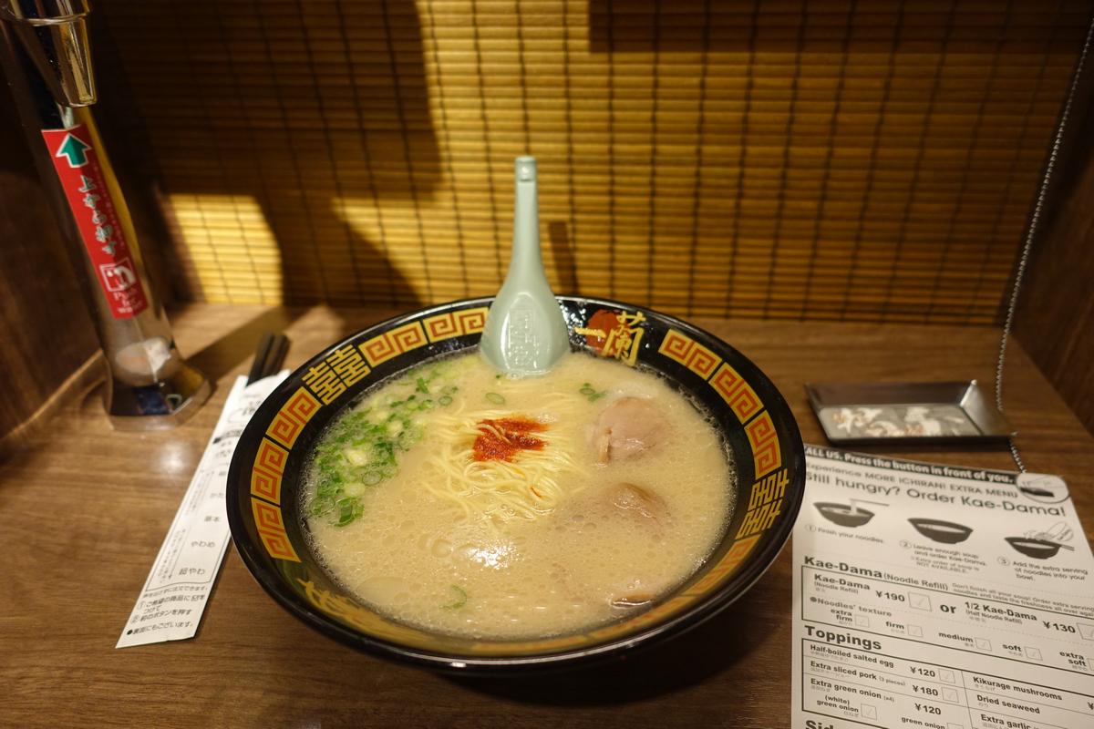A bowl of Ichiran ramen with a spoon, chopsticks, and menu is shown on a wooden counter.