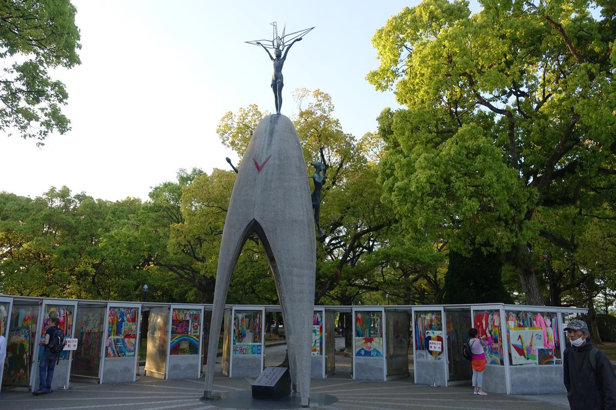 The Children's Peace Monument with a child statue holding a crane, surrounded by colorful paper cranes and green trees at Atomic Peace Park in Hiroshima.