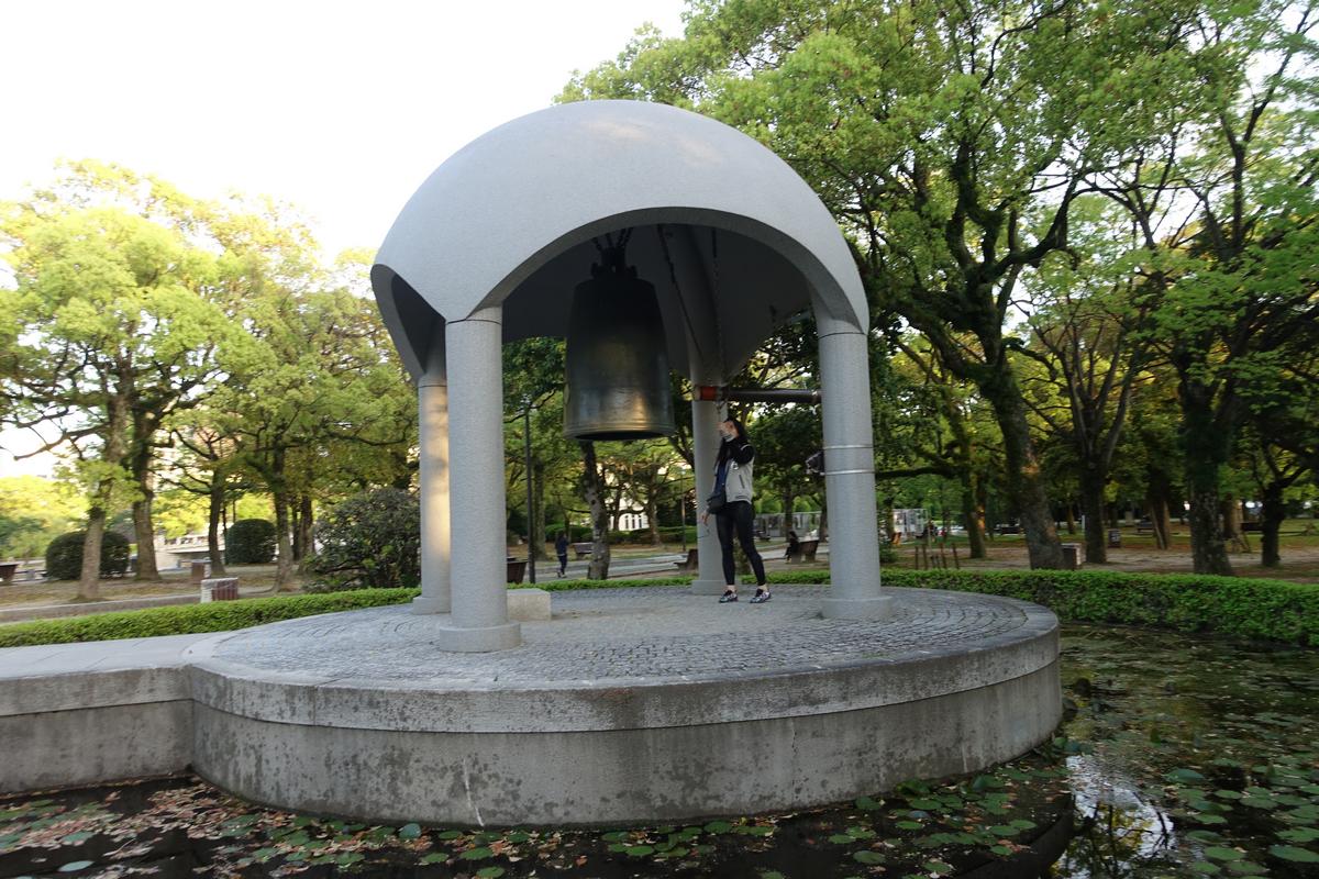 A visitor stands by the Peace Bell monument in Hiroshima's Atomic Peace Park, surrounded by a pond and green trees.