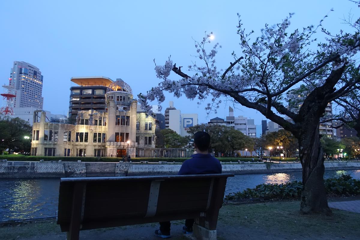 A person on a bench views the illuminated Atomic Bomb Dome and cherry blossoms in Hiroshima Peace Park at twilight.