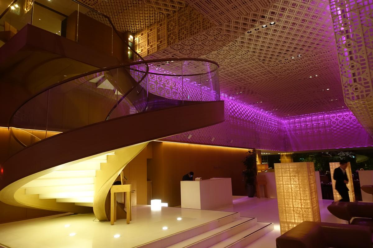 A stylish hotel lobby featuring a grand spiral staircase and a unique patterned ceiling illuminated by purple lighting.