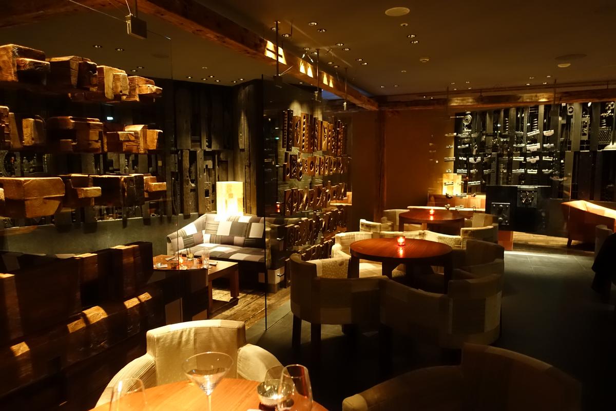 An atmospheric, dimly lit bar with dark wood decor, plush seating, and warm candlelight on tables.