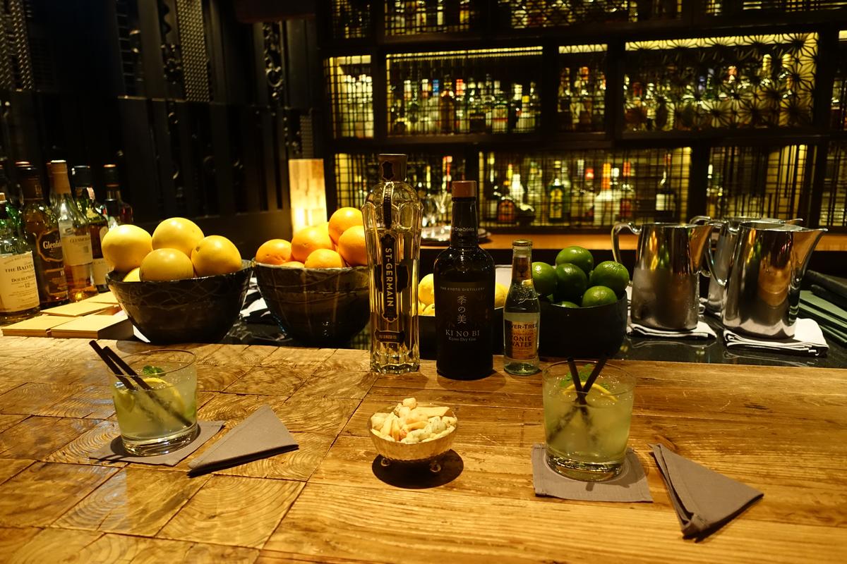 A well-stocked bar counter with two cocktails, fresh citrus fruits, and liquor bottles.