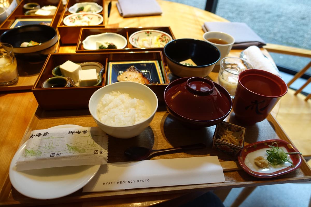 A Japanese breakfast spread with rice, grilled fish, tofu, and various small dishes arranged on a wooden table, with Hyatt Regency Kyoto branding visible.
