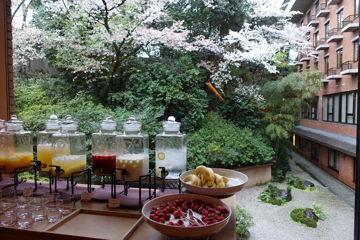 A buffet spread of drinks and fruit sits before a window overlooking a blooming cherry blossom garden and a hotel.