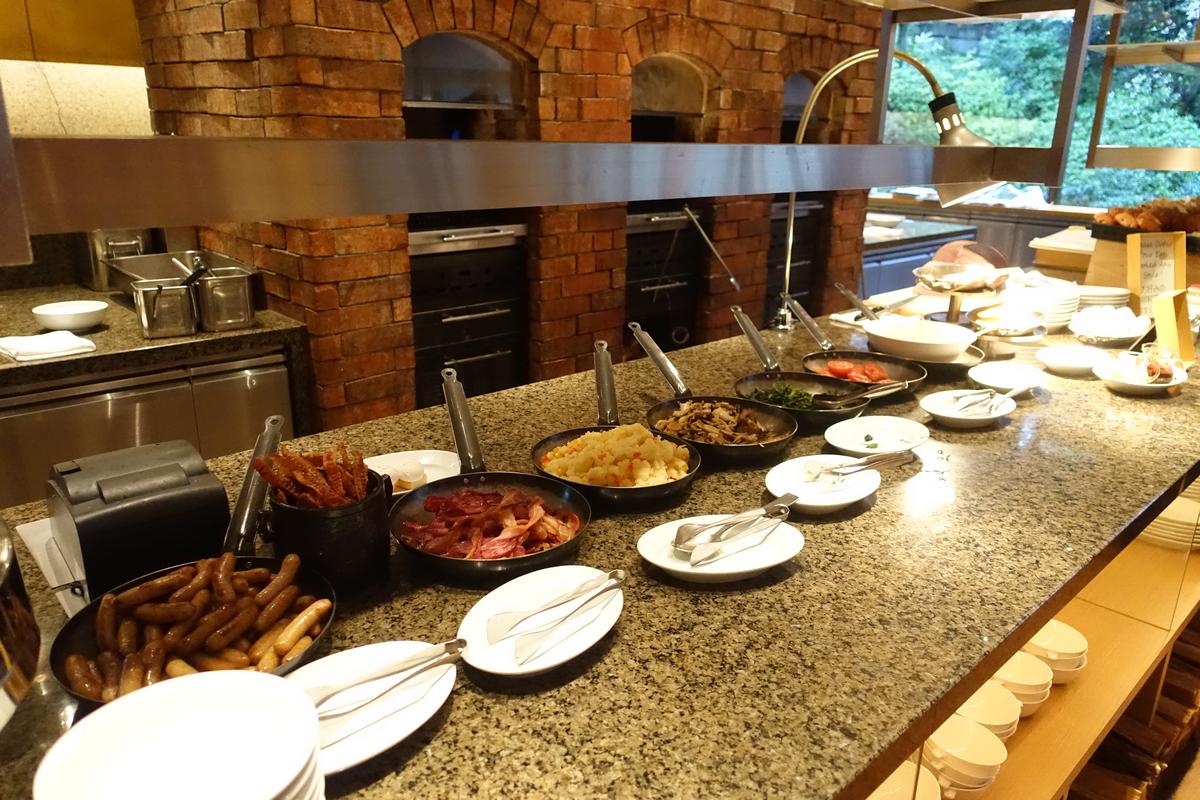 A hotel breakfast buffet features hot dishes like sausages, bacon, and scrambled eggs along a counter.
