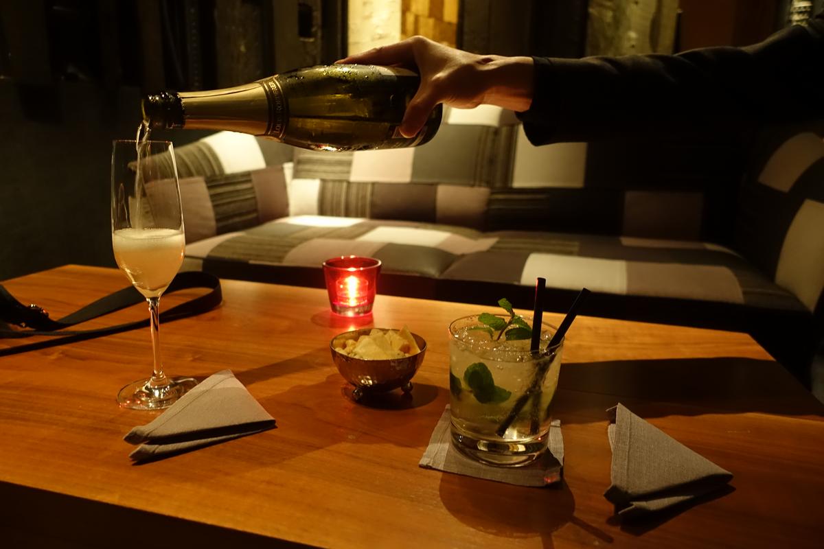 A hand pours champagne into a flute glass on a wooden table with a cocktail, snacks, and a lit candle.
