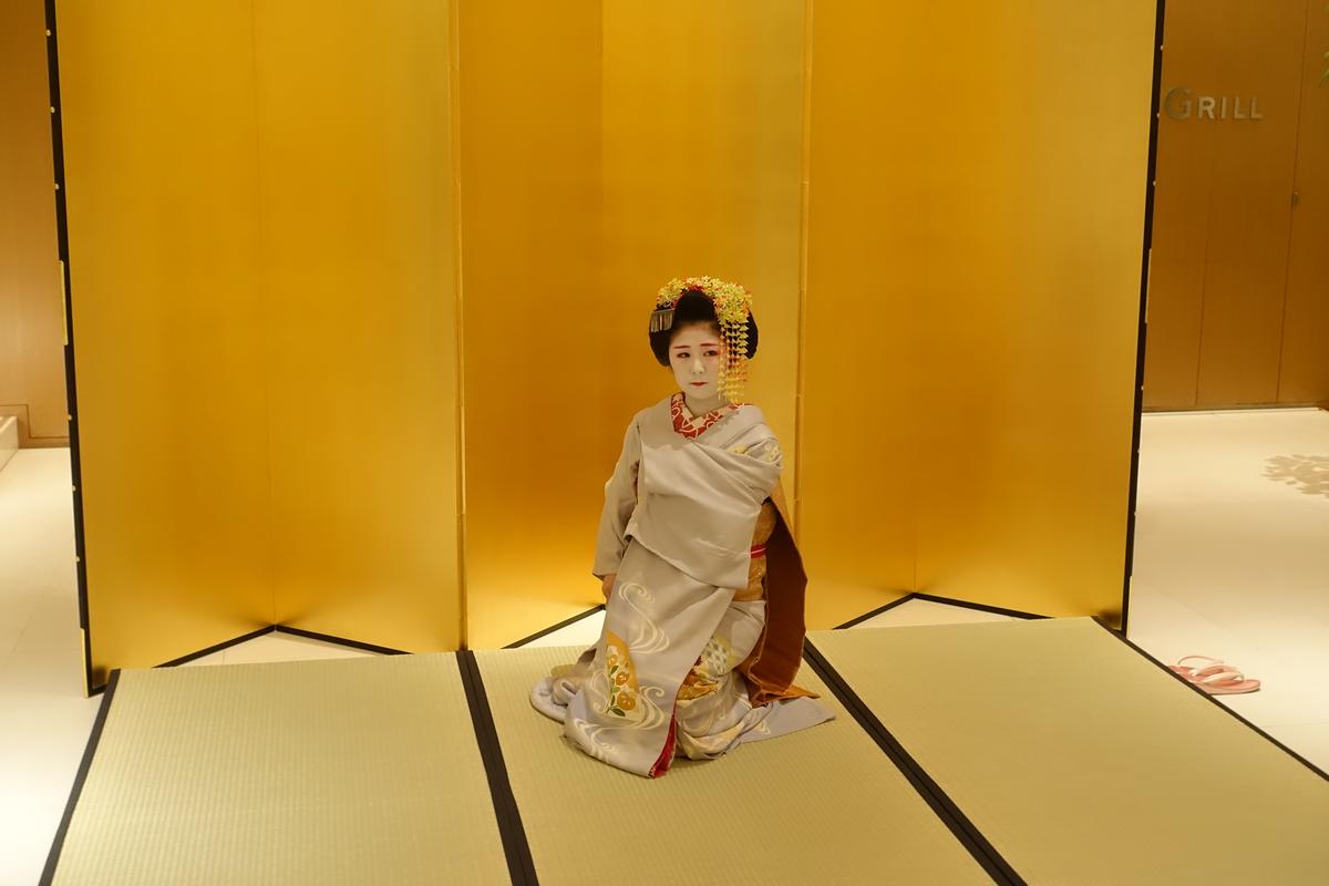A Maiko in a gray kimono kneels on a tatami mat before a golden screen.