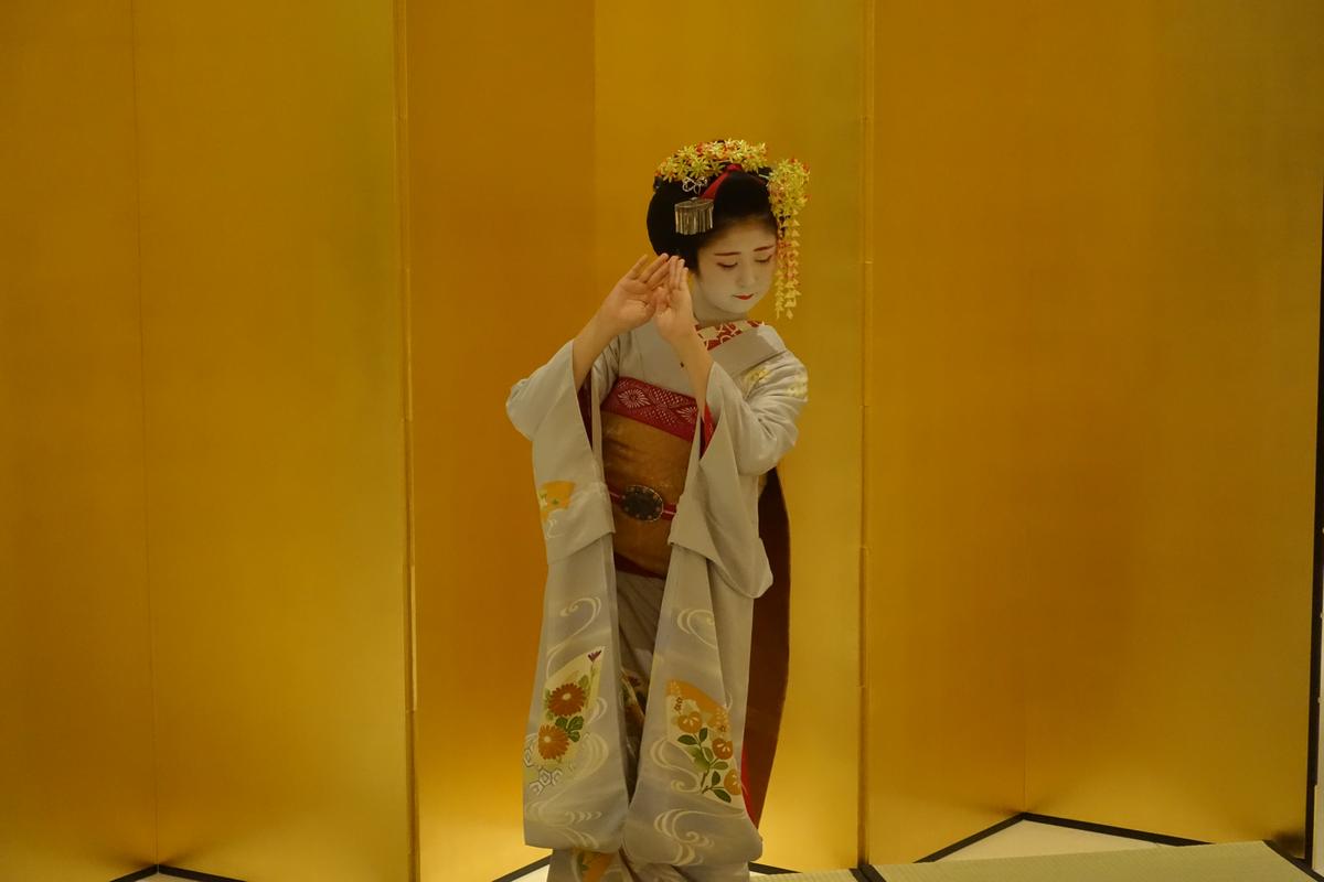 A Maiko in a light gray kimono and elaborate floral hair ornaments dances in front of a golden screen.