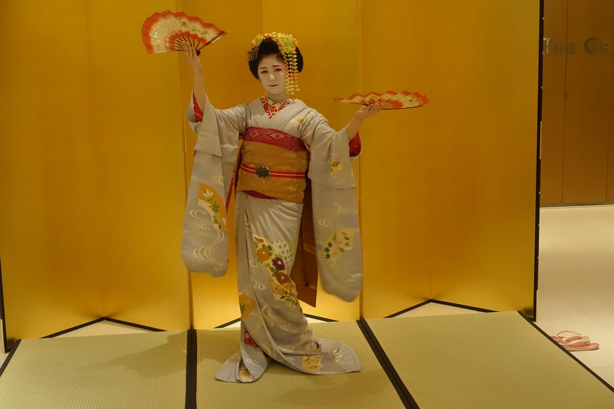 A maiko in a grey kimono with white makeup and ornate hair holds two fans while standing on tatami mats.
