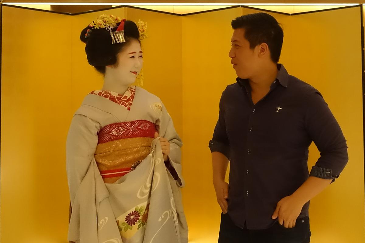 A Maiko in a traditional kimono and a man in a dark shirt look at each other and smile.
